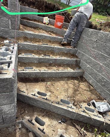 Worker building concrete steps on a stone retaining wall beside a dirt path