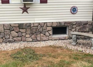 Stone-faced house exterior with a window, star decorations, a vent, and a bench beside a gravel yard.