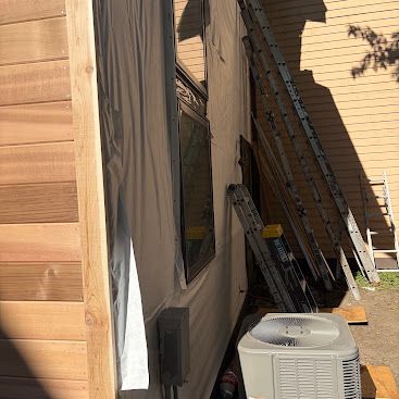 Outdoor side yard with a ladder, air conditioner unit, and stacked boards beside a tan house.