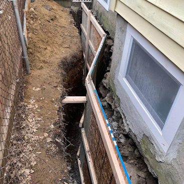 Narrow trench beside a house foundation with exposed dirt, wood supports, and a basement window.