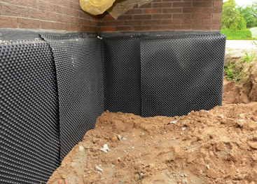 Black drainage matting on a brick foundation beside a dirt trench at a house exterior