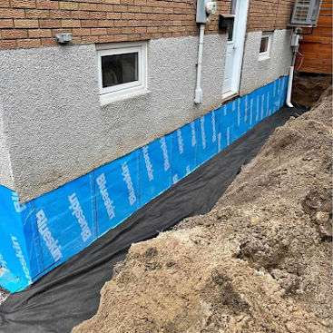 House foundation wrapped in blue waterproof membrane beside an excavated trench and dirt pile