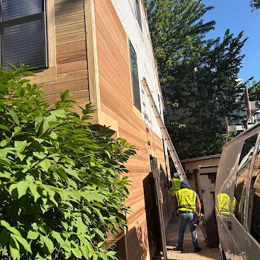 Two workers in safety vests beside a building, handling construction materials in a narrow alley.