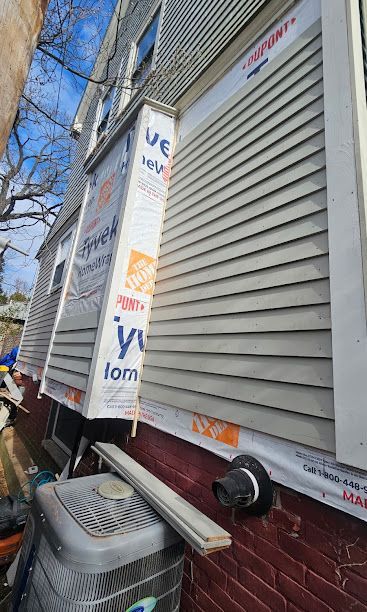 Exterior wall under renovation with white siding, red brick base, and construction wrap on a residential building