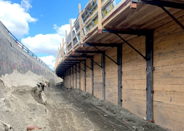 Retaining wall under construction beside an excavated dirt slope and wooden walkway under blue sky