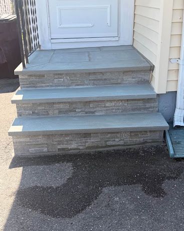 Front steps with gray tile leading to a white door, with a dark wet patch on the pavement below.