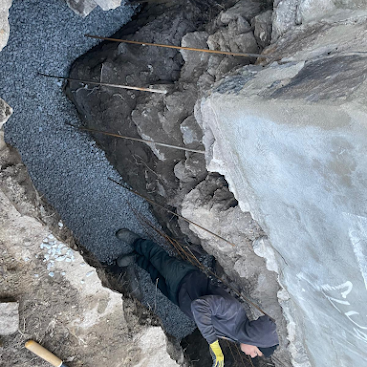 Worker crouched in a narrow rocky trench with gravel and concrete walls during excavation work