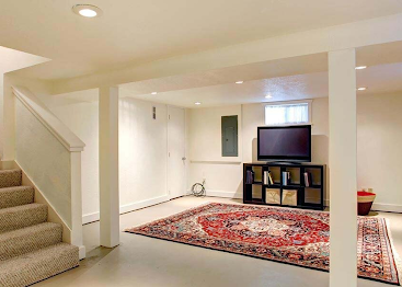 Basement living room with carpeted stairs, red area rug, TV stand, and white support columns