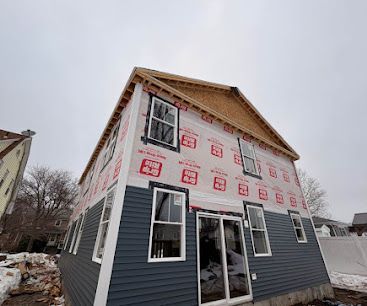 Partially finished two-story house with dark blue siding and white wrapping in a snowy neighborhood