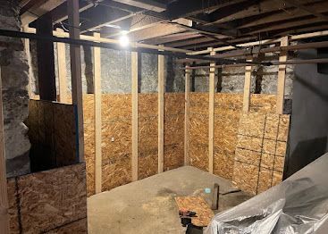 Unfinished basement with exposed framing, plywood walls, and construction materials under a single light bulb.