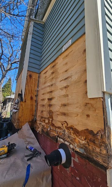Exterior house wall under repair with exposed plywood, damaged siding, and construction tools on the ground