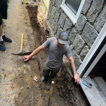 Worker digging a trench beside a stone house with a shovel and level tool on dirt ground