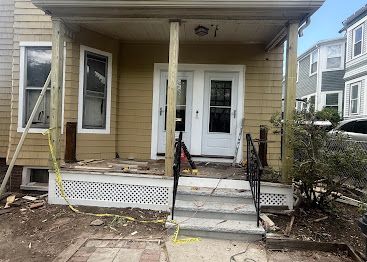Front porch of a beige house with steps, white trim, and black railings in a residential neighborhood