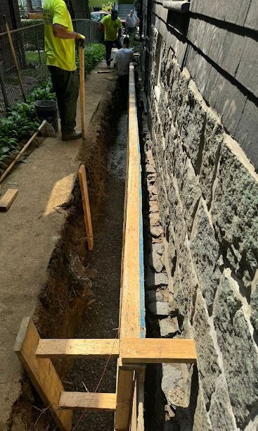 Workers dig a narrow trench beside a stone wall, with wooden supports and a wheelbarrow nearby.