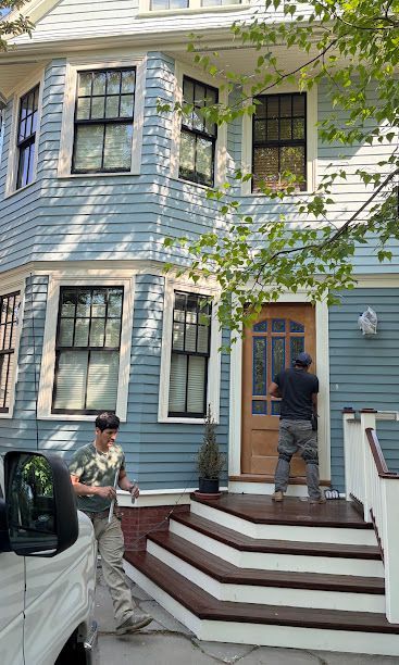 Two people near a blue house entrance, one on steps by the door and one by a parked white car.