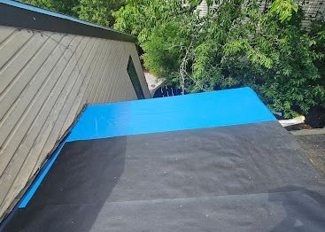Roof with black underlayment and bright blue tarp beside a green tree and beige house siding
