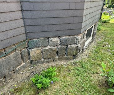 Stone foundation under gray house siding with a small basement window and patchy grass beside it
