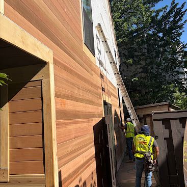 Workers on a ladder installing wood siding on a building exterior beside a utility shed