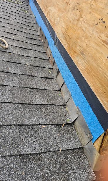 Roof shingles beside plywood wall, with blue flashing along the edge