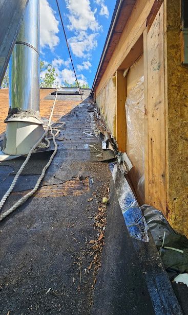 Roof view of a steep shingled roof beside a wooden wall and vent pipe under a blue sky