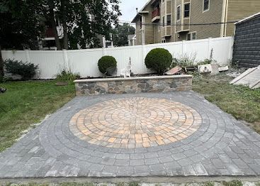 Backyard patio with gray pavers and a circular tan stone design, bordered by a small retaining wall.