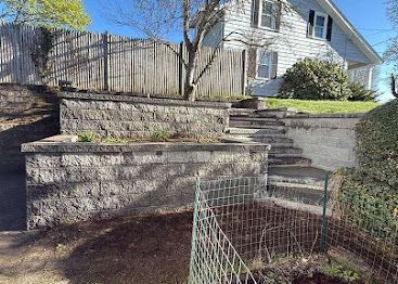 Stone retaining wall, steps, and a house on a sunny hillside with a wire fence in front.
