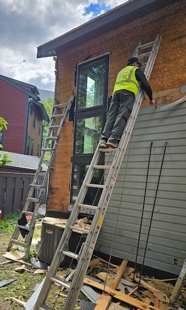 Worker in neon vest climbs a ladder on a house exterior under renovation, with tools and debris below.