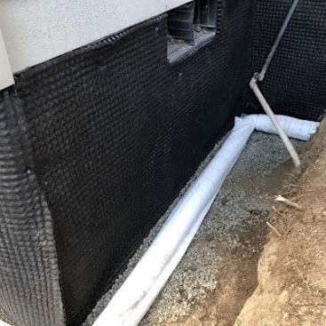 Black waterproof basement wall with white drainage pipe along gravel floor near a ground vent opening