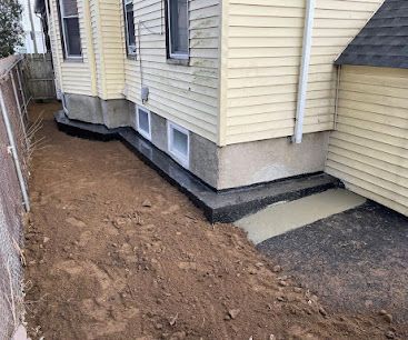 Fresh dirt trench beside a house foundation with new black waterproofing and small basement windows