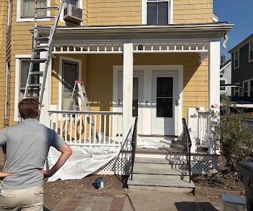 Worker standing on front porch of yellow house with ladder, likely doing exterior repairs or painting