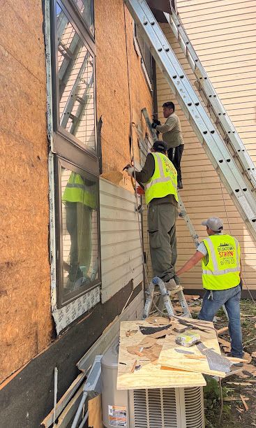 Workers install siding on a brick building beside an extension ladder and workbench.