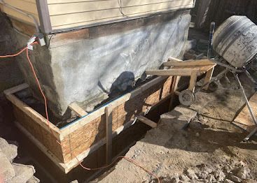 Muddy construction area with a wooden form, tarp-covered wall, and a cement mixer on the right.