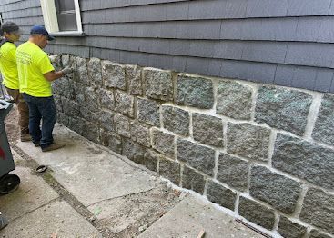 Two workers in neon shirts examining a stone foundation wall beside a house.