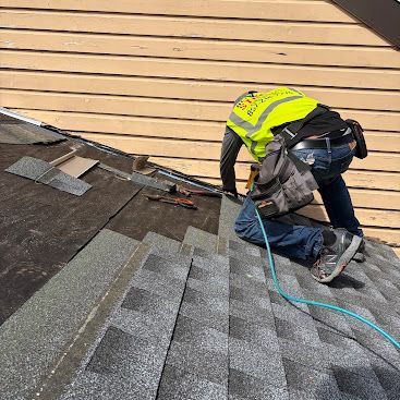 Worker installing roof shingles with a nail gun on a sloped roof, wearing a high-visibility vest.