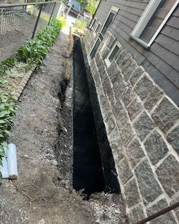 Narrow excavation trench beside a house foundation, with dirt walls and a stone-faced exterior wall.