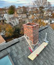 Brick chimney on a gray shingled roof, with a wooden plank and white pipe nearby.