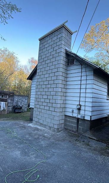 Small shingled outbuilding with a tall chimney beside a driveway under a blue sky.