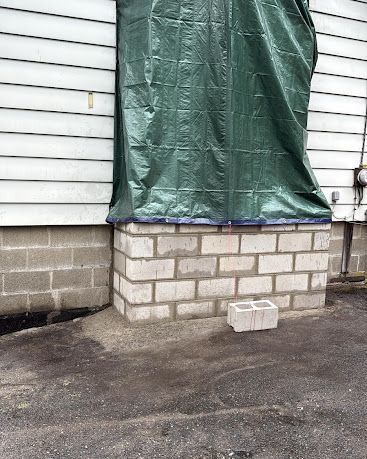 Green tarp covering a window on a white-sided house above a cinder block base, with a loose block on pavement below