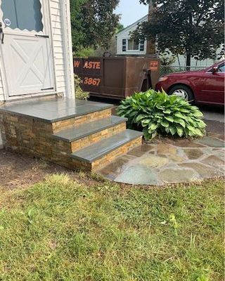 Stone steps beside a white door, with a hosta plant, stone walkway, and red car in the driveway.