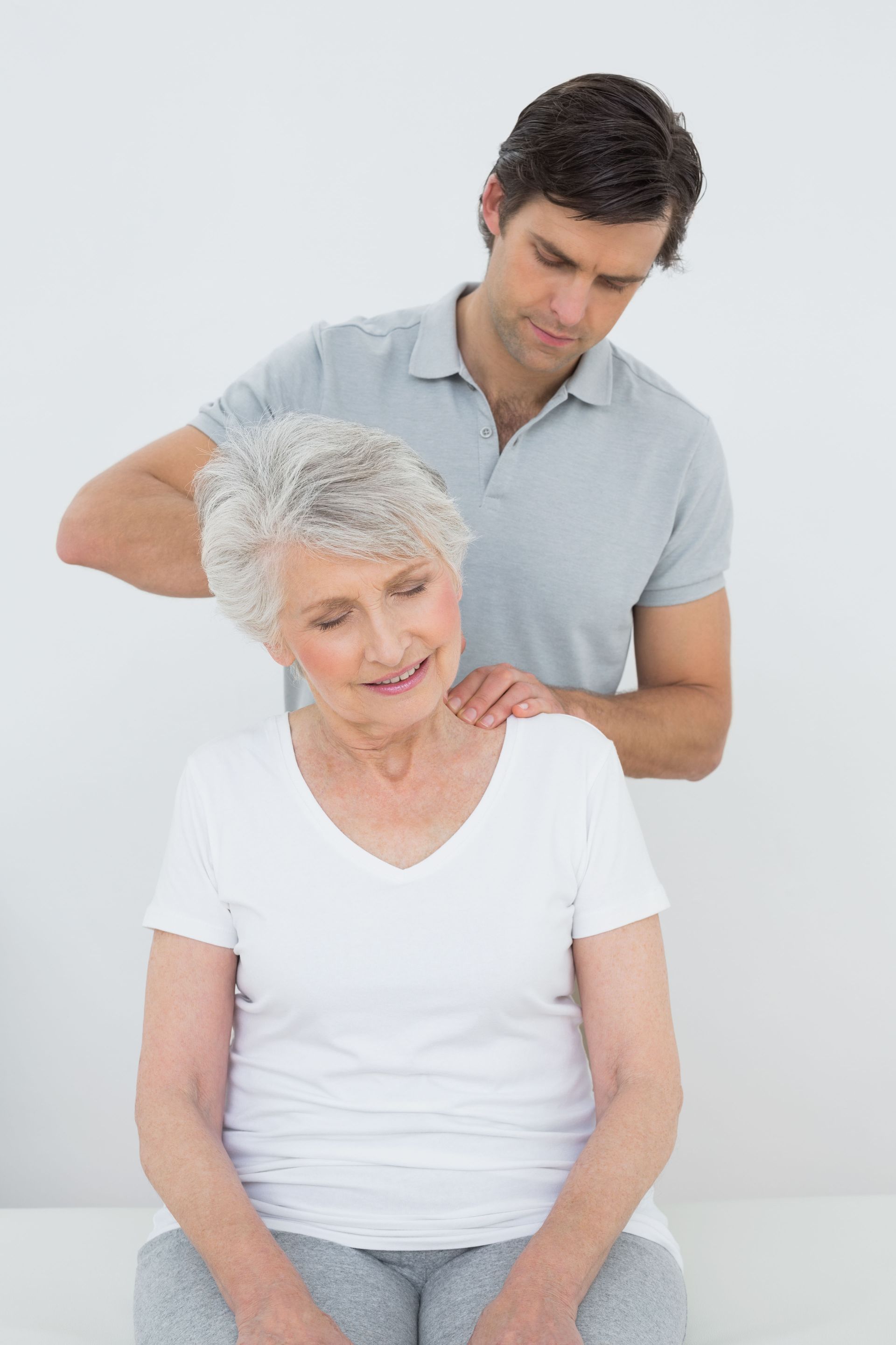Man massaging an elderly woman’s neck in a white-walled room; woman has head tilted.