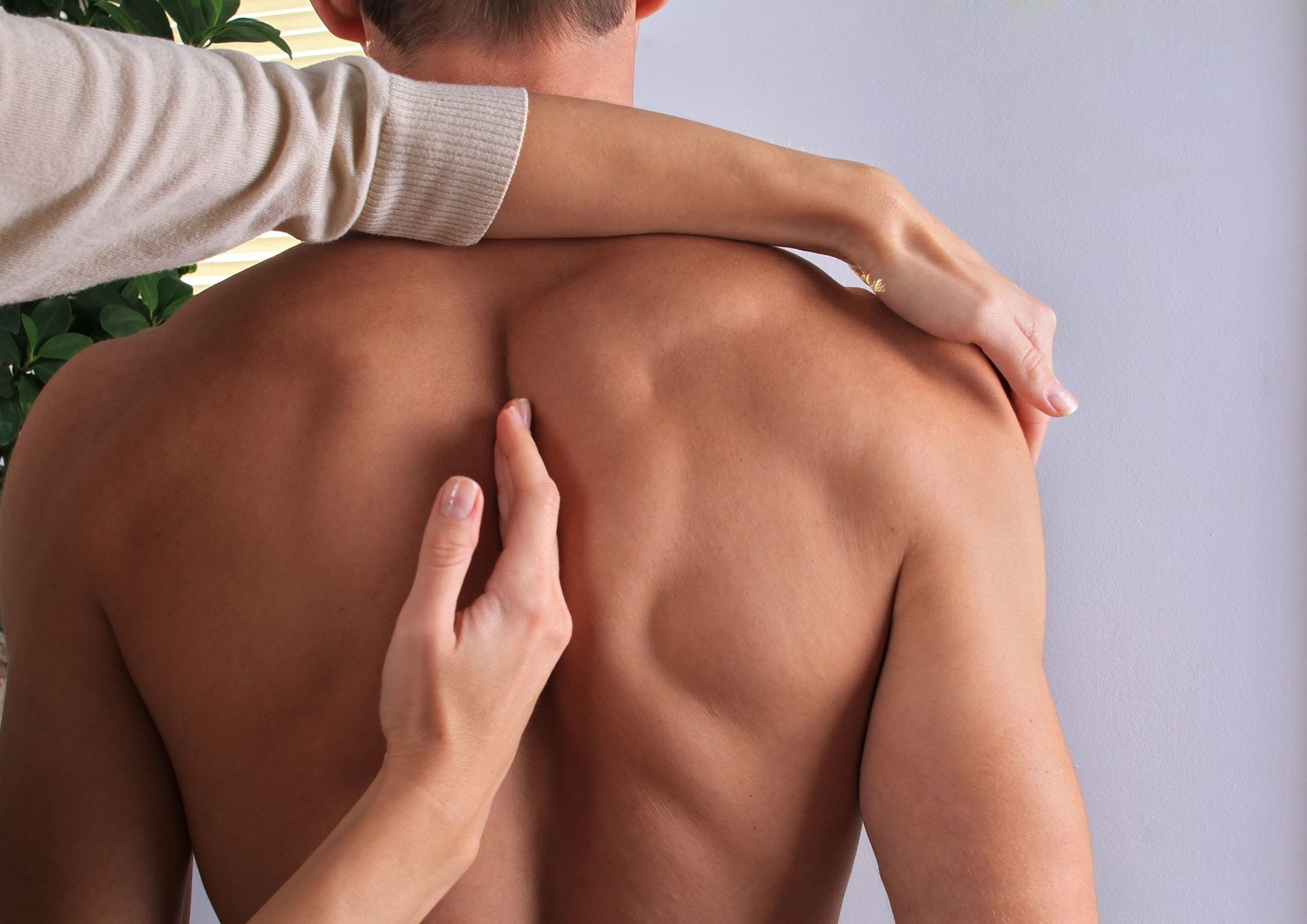 Person's bare back being examined by two hands, possibly for a medical checkup, indoors.