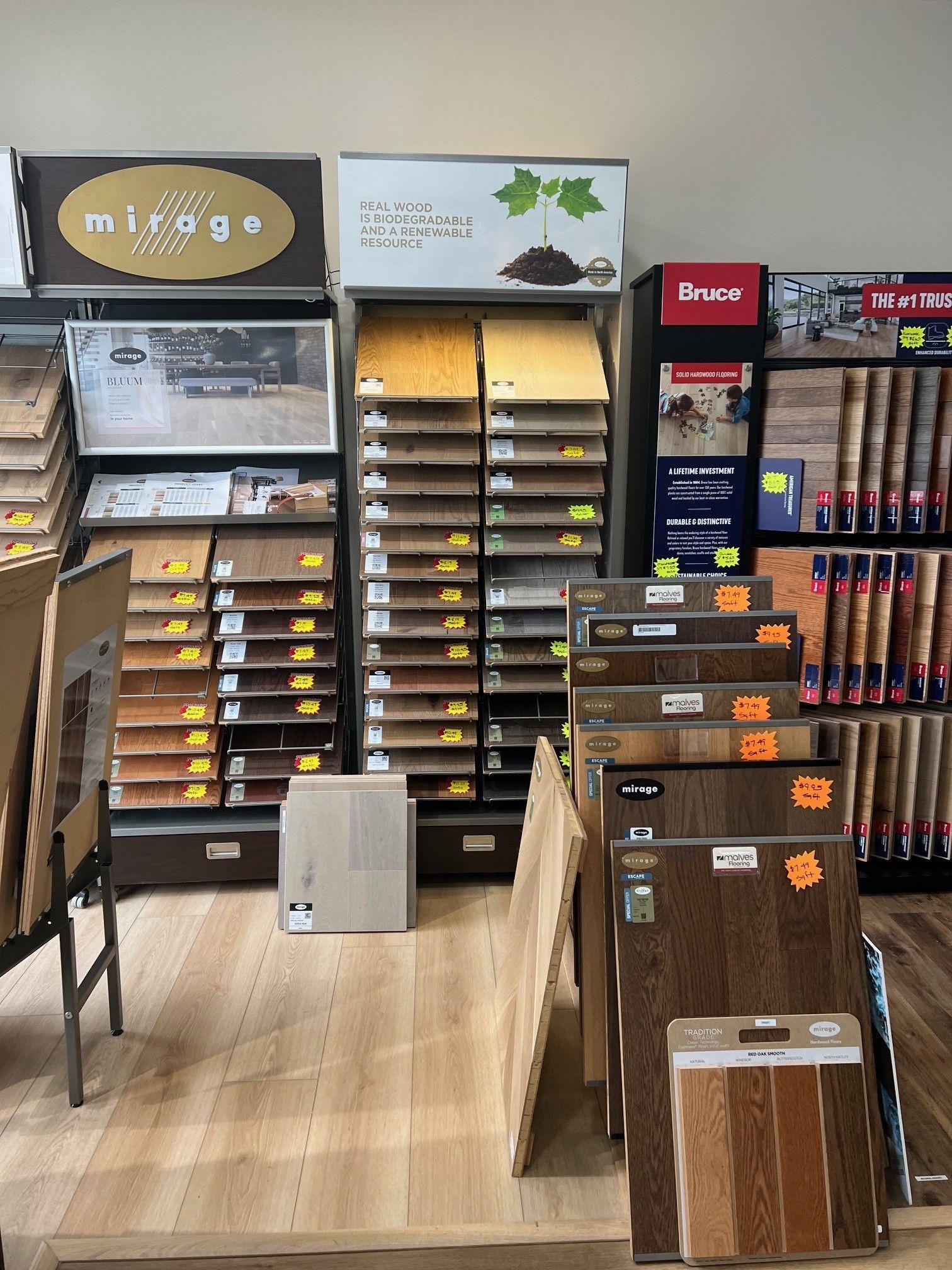 Flooring store aisle with wood and laminate samples on display racks, and boxed flooring stacks in front.