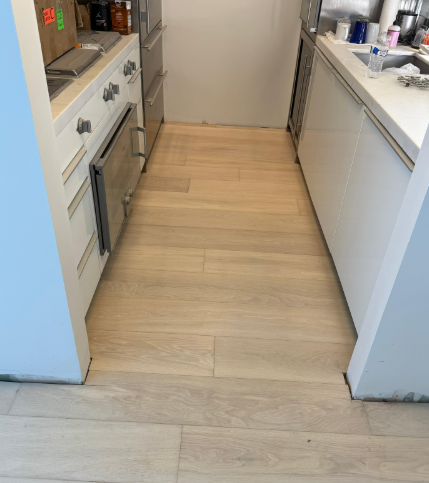 Narrow kitchen aisle with light wood flooring between white counters and cabinets, viewed toward a closed door.