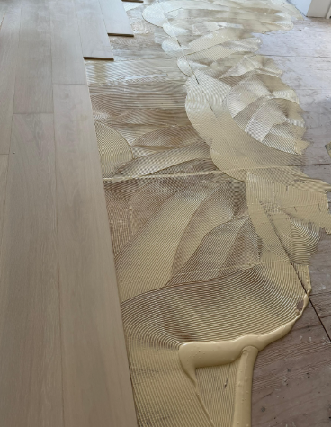 Yellow adhesive spread across a floor underlayment beside light wood flooring boards
