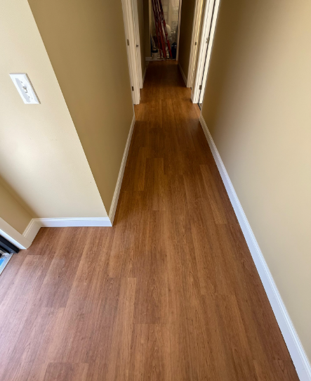 Narrow hallway with wood flooring, beige walls, and white baseboards leading to rooms in the distance