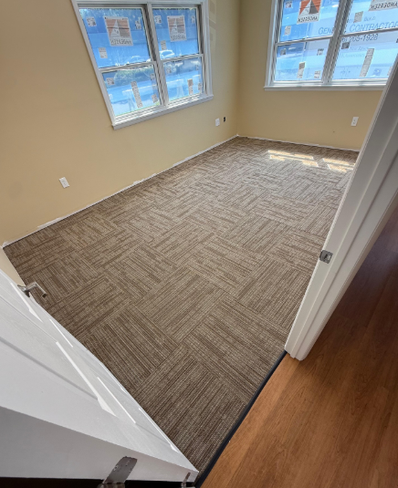Empty beige-carpeted room with two windows and a doorway, viewed from the hall.