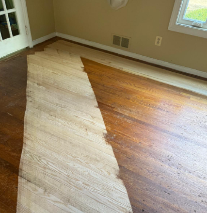 Partially sanded hardwood floor with a diagonal strip of exposed wood in a room with beige walls and windows