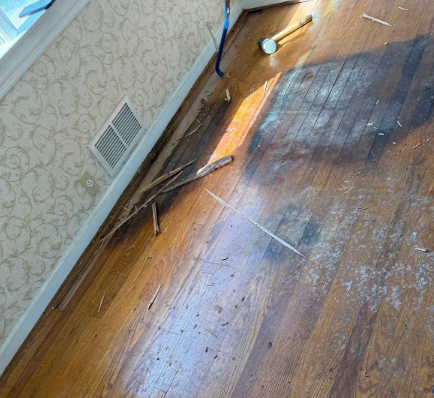 Damaged wooden floor with broken boards, debris, and a wall vent in a room.