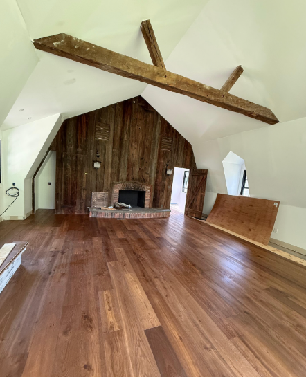 Empty loft room with wood floors, exposed beams, and a brick fireplace on a wood-paneled wall.