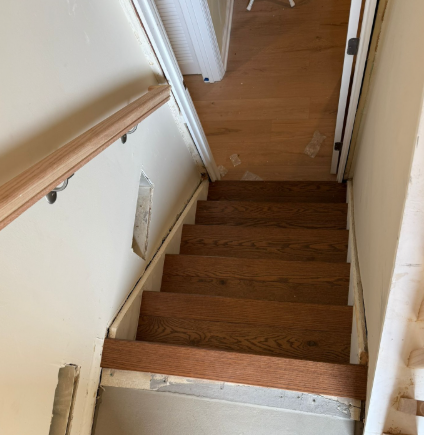 Wooden staircase viewed from the top, with white walls and a handrail in a narrow hallway.